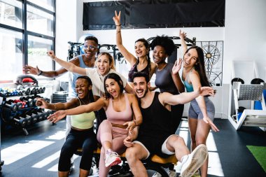 Antrenmandan sonra kollarını kaldırarak çağdaş spor salonunda toplanırken aktif giyimli, mutlu bir şekilde gülümseyen bir grup pozitif genç kadın ve erkek.
