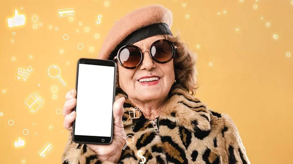 Content older woman showing cellphone with empty screen against yellow backdrop with icons of shopping trolley and gifts near dollar symbol and like button