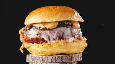 İştah açıcı hamburgerin peynir ve mayonezin altında kızartılmış çöreklerle siyah arka planda dönen ahşap tezgahta hareket etmesini durdurun.