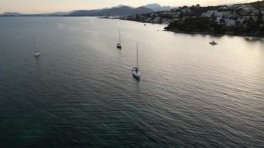 İspanya, Mallorca 'da gün batımında kıyı kasabası yakınlarındaki dalgalı deniz yüzeyinde seyreden modern yatların insansız hava aracı görüntüsü inanılmaz.