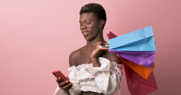 非洲变性人在一个孤立的背景下拿着购物袋 一边用手机一边笑 技术和采购概念 — 图库视频影像