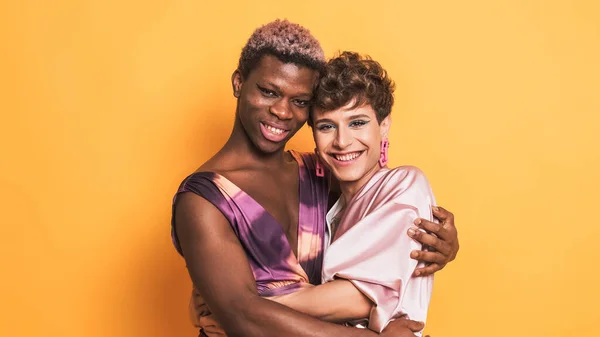 two drag men couple in make up and fashion clothes embracing and smiling together looking to camera on yellow background