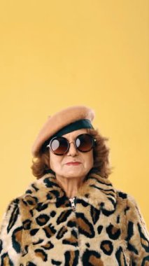 Stylish senior woman in sunglasses wearing a leopard fur coat and a beret while smiling over an yellow background.