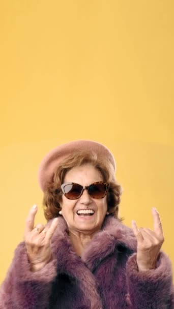 站在一个黄色的背景上 一个时髦的老年妇女用手指做着一个摇滚动作 — 图库视频影像