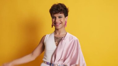 Pembe elbiseli transseksüel bir adamın videosu ve sarı arka planda kameraya gülümseyen bir makyaj.