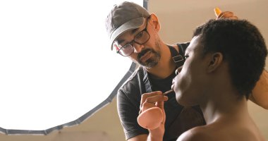 Düşük açılı odaklanmış sakallı, gözlüklü, dudak parlatıcısı sürerken stüdyodaki travesti modeline makyaj yapıyor.