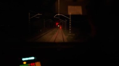 Karanlık bir gecede, farlı ve boş tren rayı ile elektrik direklerinin yakınındaki kırmızı bir durağa giden tren.
