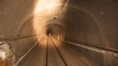 Çakıl taşı yüzeyindeki boş raylar trenin farlarını yakarken aydınlatılmış kemer tünelinden geçiyor ve turuncu sinyalle yan taraftaki elektrik direklerine yaklaşıyor.