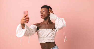 Kadın elbiseli Afro-Amerikan transseksüel modeli pembe arka planlı fotoğraf stüdyosunda telefonla selfie çekiyor.
