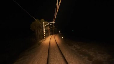 Gerçek zamanlı çalışan trenler ve boş raylar, çakıl yüzeyinde, tedavi edilmiş yolda, elektrik direkleriyle, yeşil ağaçların yanında, yeşil ışığa yaklaşan elektrik direkleriyle, karanlık gecede.