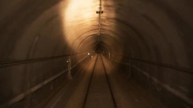Çakıl taşı yüzeyindeki boş raylar trenin farlarını yakarken aydınlatılmış kemer tünelinden geçiyor ve turuncu sinyalle yan taraftaki elektrik direklerine yaklaşıyor.