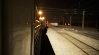 Yansıyan tırmıklarla gerçek zamanlı tren manzarası. Yavaş hareket ediyor ve aydınlık tren istasyonuna giriyor. Platformda duruyor ve gece vakti kırmızı ışıkta geçiyor.