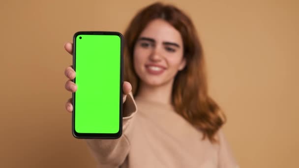 ベージュの背景に立っている緑のスクリーンとスマートフォンを示しながら カメラを見ている暗い髪のカジュアルな服で幸せな若い女性 — ストック動画