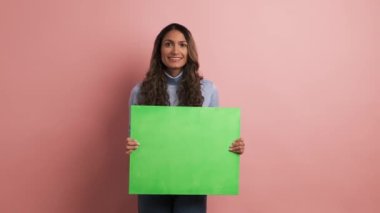 Pembe arka planda elinde yeşil bir tahta tutan ve baş parmağını kaldıran gülümseyen bir kadın videosu.