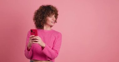 Pembe elbiseli, kıvırcık saçlı, akıllı telefonlu gülümseyen genç bir kadın pembe arka planı işaret ederken...