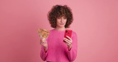 Pembe elbiseli, kıvırcık saçlı, elinde para tutan ve pembe arka plana karşı akıllı telefon kazanan genç bir kadın.
