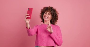 Pembe elbiseli, kıvırcık saçlı, gülümseyen genç bir kadın. Pembe arka planda durarak akıllı telefonuyla selfie çekerken dudaklarını büzüyor.