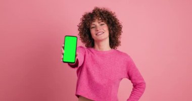 Kıvırcık saçlı gülümseyen kadın pembe arka planda yeşil ekranlı akıllı telefon gösteriyor.
