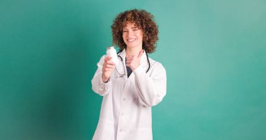 Gülümseyen kıvırcık saçlı kadın doktor yeşil arka plana karşı baş parmağıyla ilaç şişesi uzatırken kameraya bakıyor ve hareket ediyor.