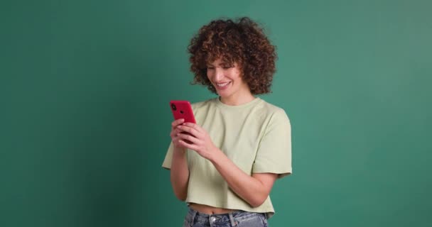 Glimlachende Jonge Vrouw Met Krullend Haar Met Mobiele Telefoon Tegen — Stockvideo