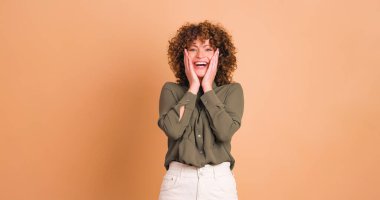 Heyecanlı, kıvırcık saçlı, makyajlı, kolları dolu genç bir kadın kameraya bakıyor, el çırpıyor ve bej arka planda başarıyı kutluyor.