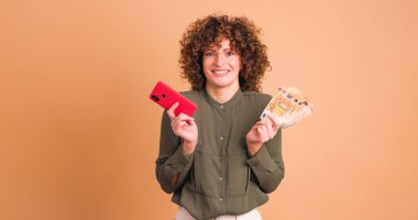 Pembe elbiseli, kıvırcık saçlı, elinde para olan gülümseyen genç bir kadın ve bej arka plana karşı akıllı telefon kazanan piyango kullanıyor.