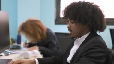Afro saç stili olan, masa başında bilgisayar kullanan ve ortak çalışma alanındaki kameraya bakan mutlu, genç, Afrikalı, yaratıcı iş adamı.