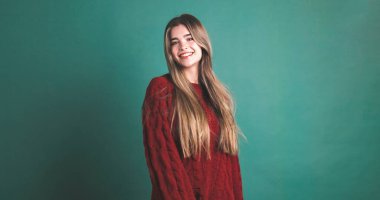 Kestane rengi kazaklı, uzun sarı saçlı ve yeşil arka planda kameraya bakan gülümseyen genç bayan model.