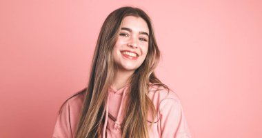 Stüdyoda şeftali rengi arka planda dikilirken gülümseyen ve kameraya bakan şeftali rengi, uzun saçlı kadın portresi.