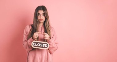 Pembe kapüşonlu, uzun sarı saçlı, elinde pembe arka planda kapalı levhayı tutarken kameraya bakan üzgün genç bayan.