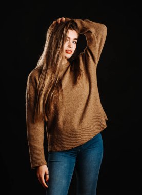 Kahverengi kazaklı, uzun sarı saçlı, siyah arka planda dikilirken kameraya bakan genç ve şehvetli kadın modelin portresi.