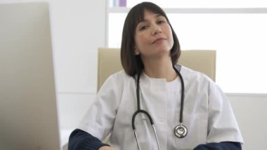 Kendine güvenen genç bayan doktor, klinikte sandalyeye oturmuş steteskopla kameraya bakarken yazıp gülümsüyor.