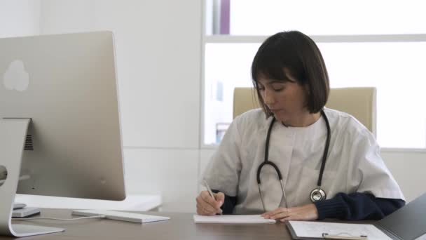 集中的女医生 脖子上挂着听诊器 坐在诊所的办公室里写信息和浏览台式计算机 — 图库视频影像