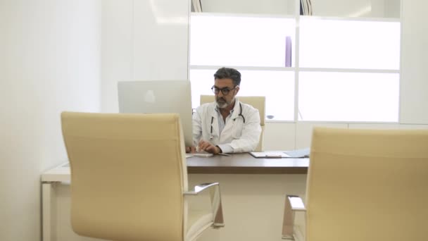 在现代诊疗所的工作天 用计算机实时放大集中 头发灰白 脖子上有听诊器的成熟男医生 然后在工作天做笔记 — 图库视频影像