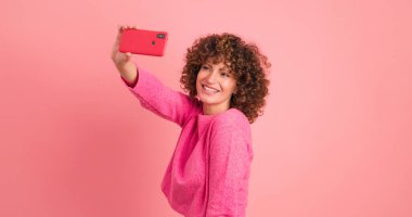 Pembe elbiseli, kıvırcık saçlı, gülümseyen genç bir kadın pembe arka planda duran akıllı telefonuyla selfie çekiyor.