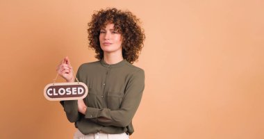 Elinde kapalı levhası olan kıvırcık saçlı kadın, bej arka planda üzgün..