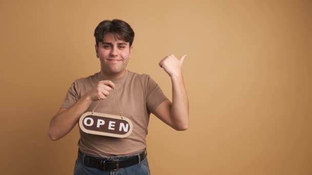 ベージュの背景に歓迎しながら開いたサインを保持するカジュアルな服でヒスパニックの若い男性を微笑む肖像画 — ストック動画