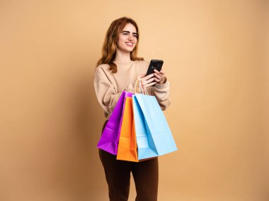 Koyu renk saçlı, günlük kıyafetlerle gülümseyen genç bir kadın. Renkli alışveriş poşetleri taşırken akıllı telefon kullanıyor. Bej arka planda dikilirken başka tarafa bakıyor.