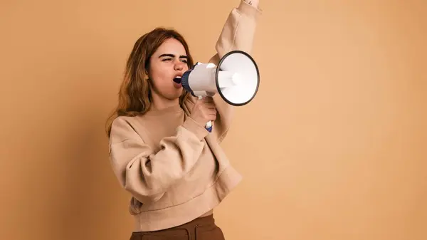 Young female activist in casual clothes with dark hair shouting on megaphone while protesting against beige background