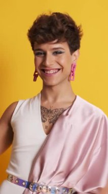 Pembe elbiseli bir transseksüelin dikey videosu ve sarı arka planda kameraya gülümseyen bir makyaj malzemesi.