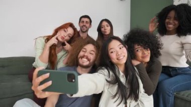 Koltukta ve yerde oturan mutlu çok ırklı arkadaşların yavaş çekimleri cep telefonuyla selfie çekerken iki parmağını hareket ettiriyor ve gülümsüyor.