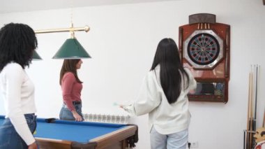 Çeşitli arkadaşlar dart tahtasının hedefine dart okları atan ve hafta sonu evdeki oyun odasında başarının tadını çıkaran kadınları seyrediyor.