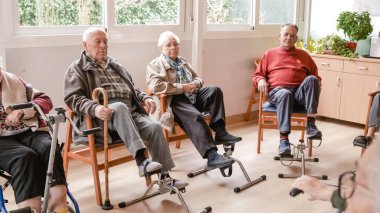 Yaşlı erkekler ve kadınlar, rehabilitasyon merkezindeki fizik tedavi seansları sırasında tahta koltuklarda oturup pedal egzersizleri yapıyorlar.