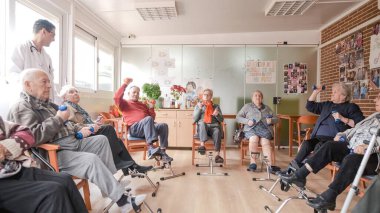Rehabilitasyon merkezindeki fizyoterapi seansında pedal egzersizi yapan ve dambılları kaldıran yaşlı erkekleri izleyen mutlu erkek bakıcı.