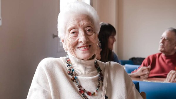 Portrait of elderly woman with short gray hair adorned with necklace smiling directly at camera while seated in nursing home