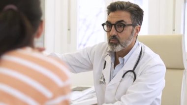 Üniformalı neşeli, olgun bir doktor. Steteskop boynunda. Masada bilgisayarla oturuyor ve kadın hastalarla konuşuyor.