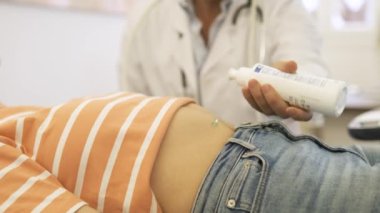 Üniformalı bir erkek doktor, kadın hastanın karnına jel dökerek kanepede yatar sonra da ultrason yapar.