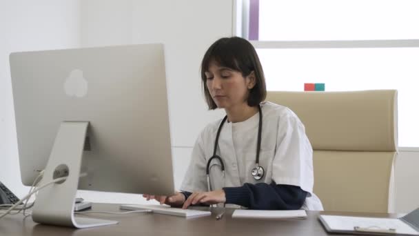 集中精神的女医生 脖子上挂着听诊器 坐在诊所的办公室里写信息和浏览台式计算机 — 图库视频影像