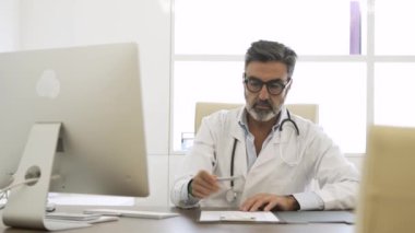 Olgun erkek doktor panoya iliştirilmiş bir şeyi not alıyor ve klinikteki masada bilgisayar kullanıyor.
