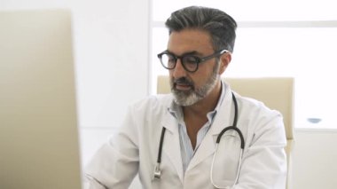 Steteskobu boynuna dolamış kendinden emin yetişkin bir erkek doktor. Klinikte otururken kameraya bakıyor ve konuşuyor.